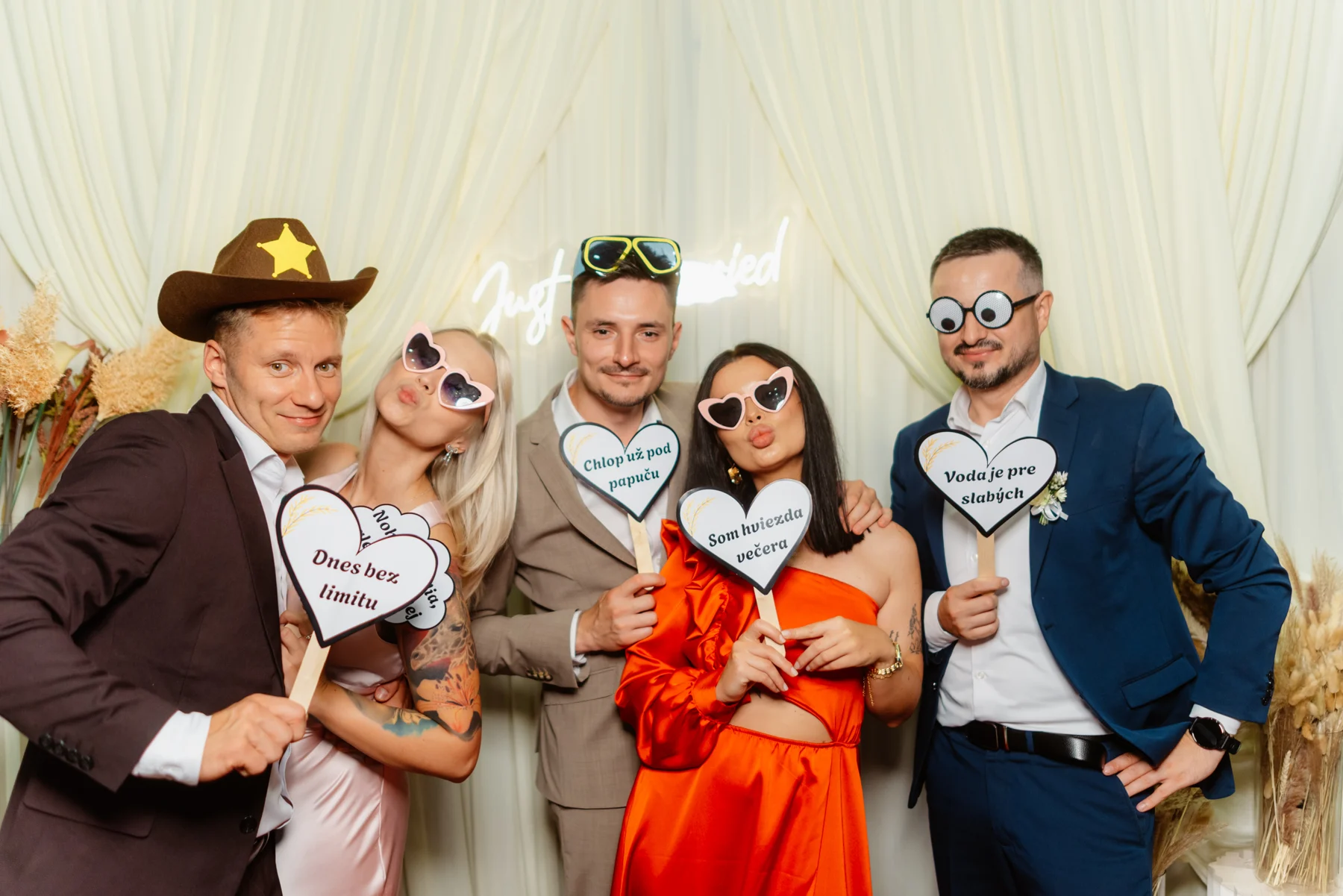 Priatelia pózujú pred fotoboxom s klobúkmi, okuliarmi a tabuľkami