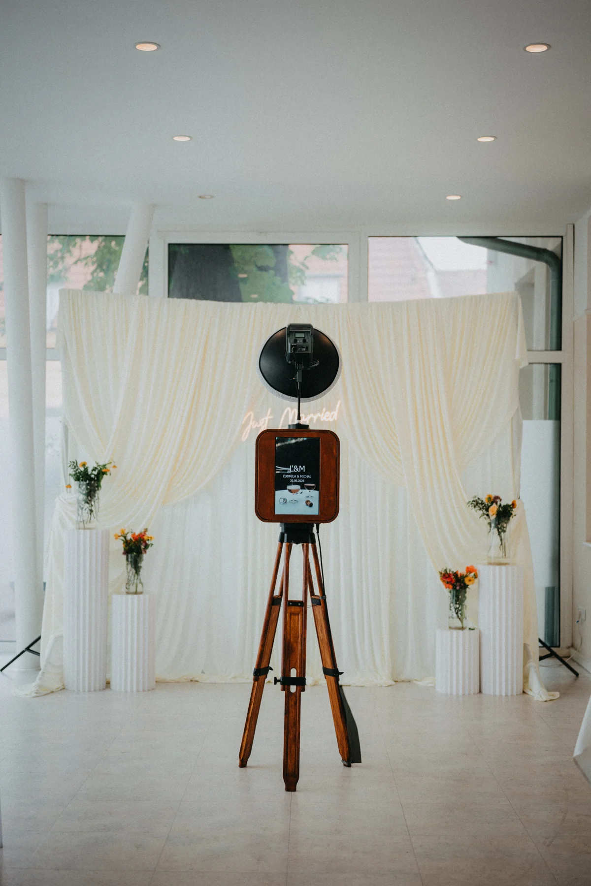 Drevený fotobox na statíve pred bielym svadobným pozadím s kvetmi a nápisom Just Married